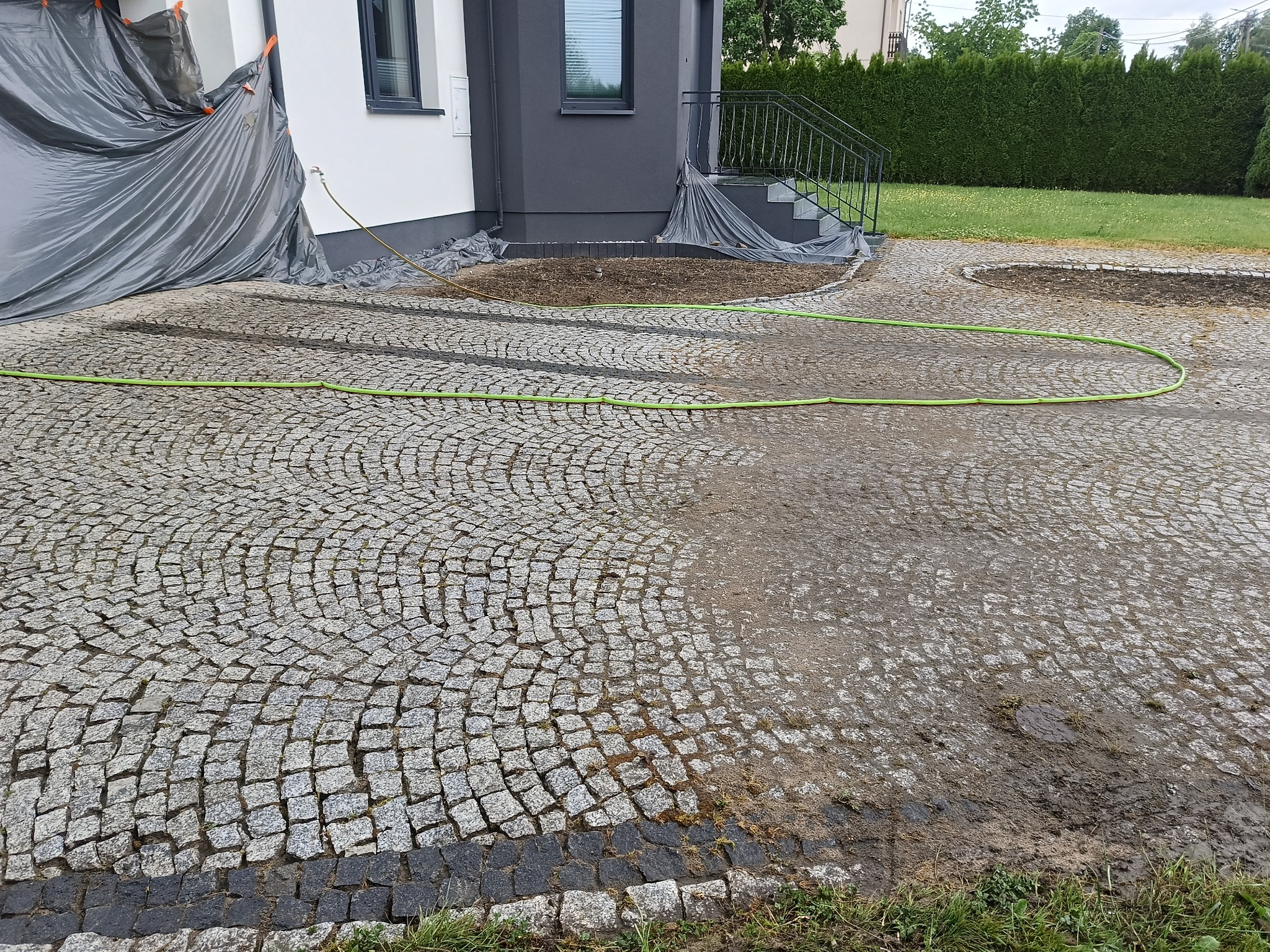 Częściowo umyta kostka brukowa na podjeździe, widoczna zielona rura doprowadzająca wodę, zabezpieczona folią elewacja budynku w tle.