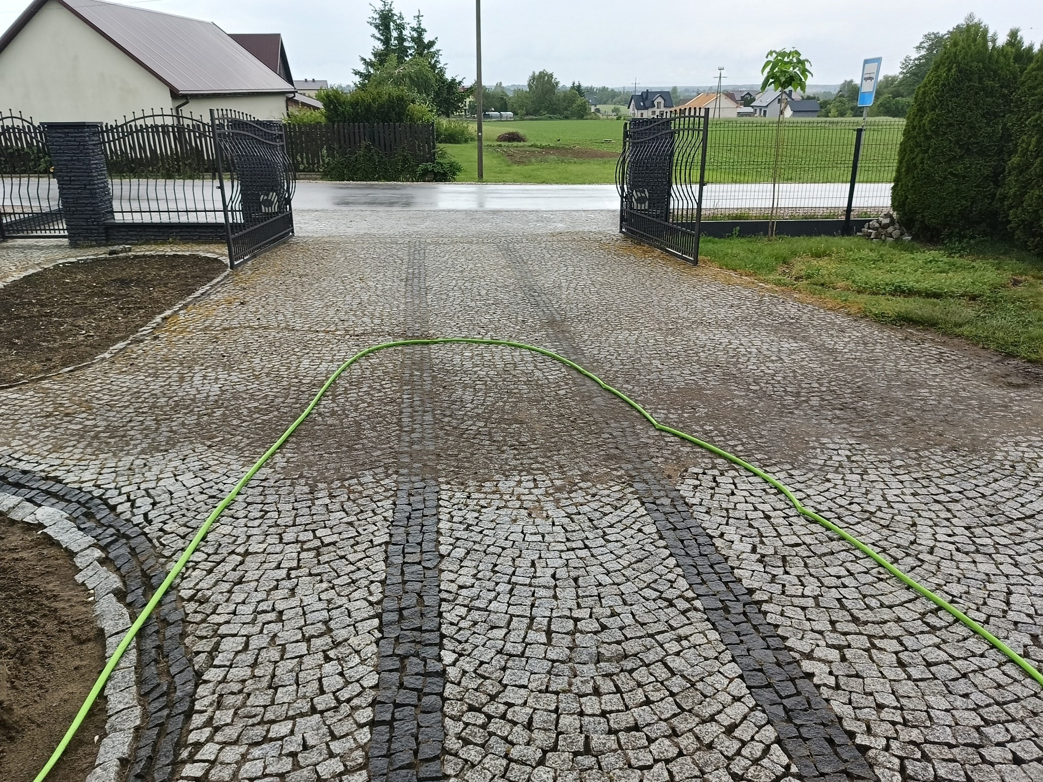 Mokra kostka brukowa na podjeździe z otwartą bramą, widoczne ślady po umyciu i zielony wąż ogrodowy