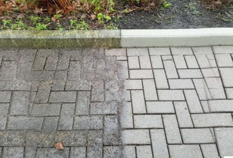 Częściowo oczyszczona, szara kostka brukowa ułożona we wzór jodełki, widoczna różnica w kolorze i czystości między umytą a brudną powierzchnią, obok rabata z roślinami.