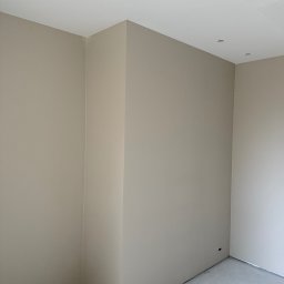 Adrian Nowak - Wnętrze po malowaniu ścian na beżowo-szary odcień, widoczne gładkie powierzchnie i otwory na oświetlenie w suficie. Podłoga betonowa, sufit biały z perforacją.