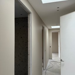 Adrian Nowak - Jasny korytarz w trakcie malowania, widoczne surowe ściany i ościeżnice, kabel na podłodze, światło dzienne z okna dachowego. Minimalistyczny styl.