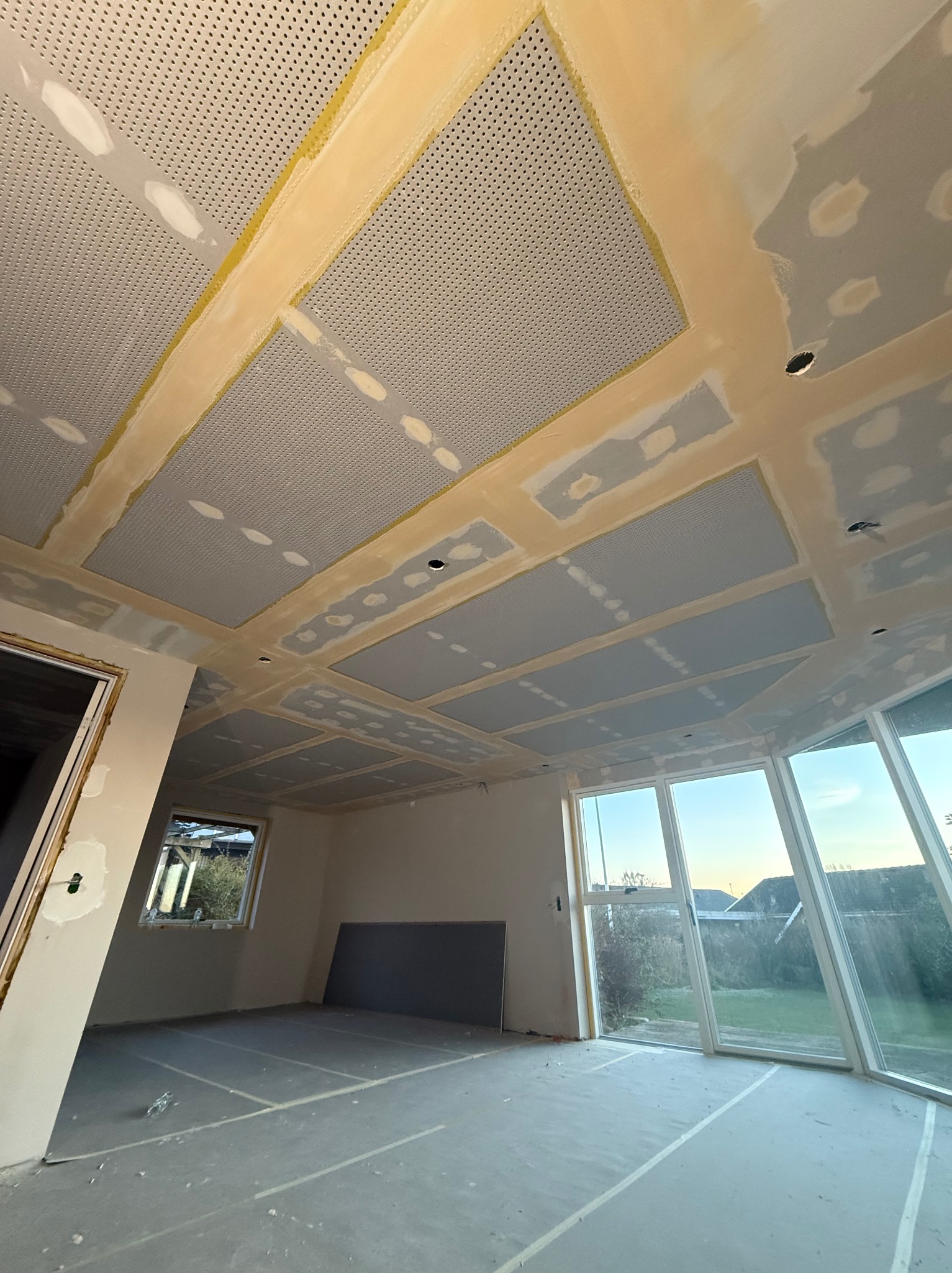Sufit z płyt kartonowo-gipsowych z perforacją, w trakcie szpachlowania łączeń, widok z dołu, duże okna w tle, przygotowanie do malowania.