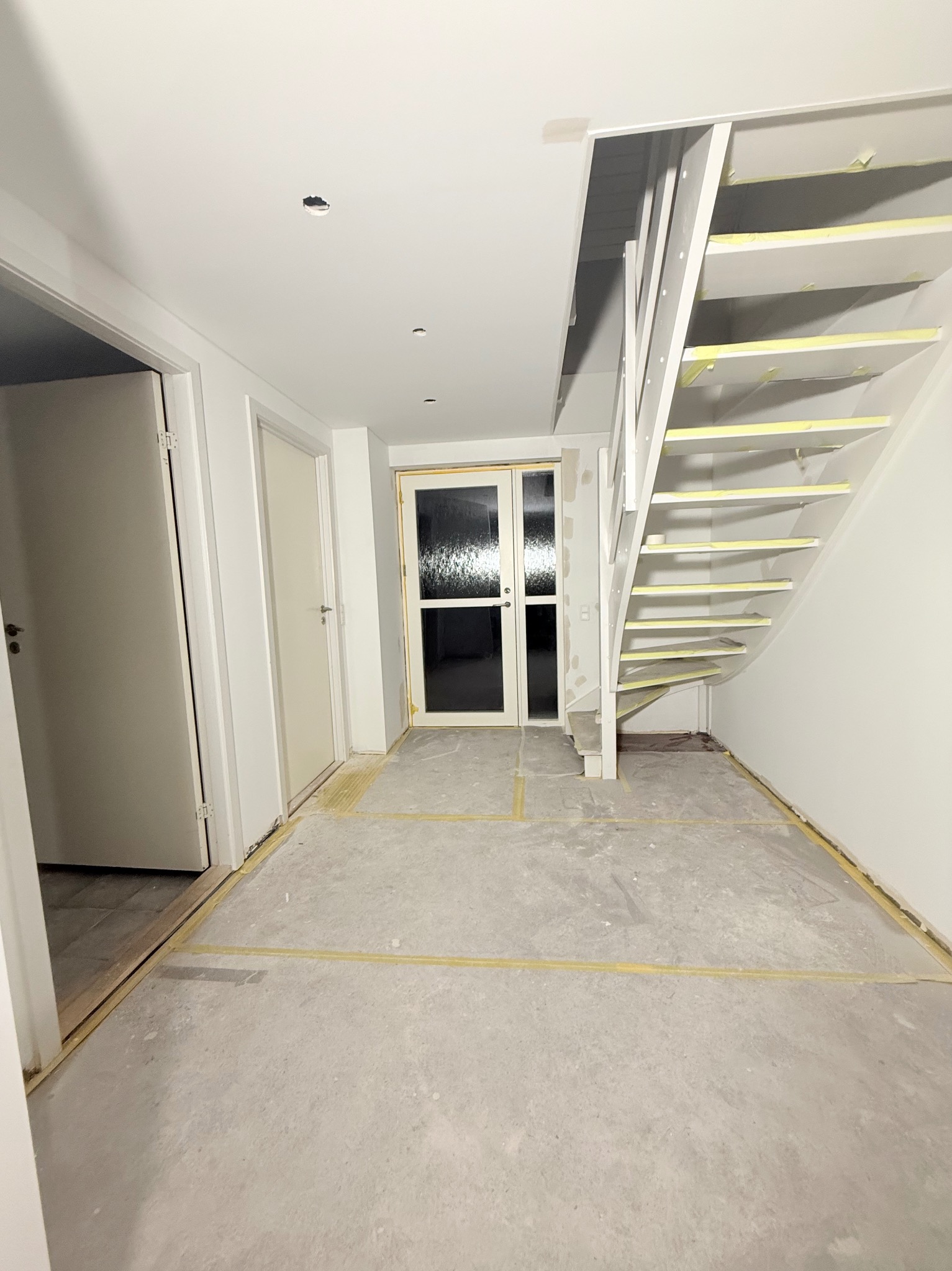 Wnętrze w trakcie remontu: białe ściany, schody zabezpieczone taśmą malarską, surowa podłoga przygotowana do dalszych prac wykończeniowych, widoczne drzwi.