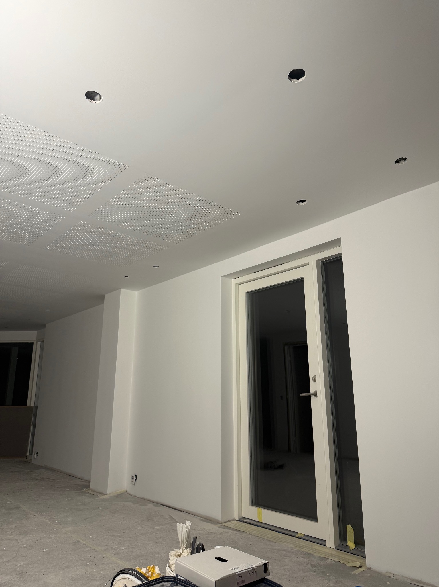Białe, świeżo pomalowane wnętrze z widocznymi otworami na oświetlenie w suficie. Na podłodze widoczne ślady po pracach malarskich, oklejone taśmą drzwi balkonowe.