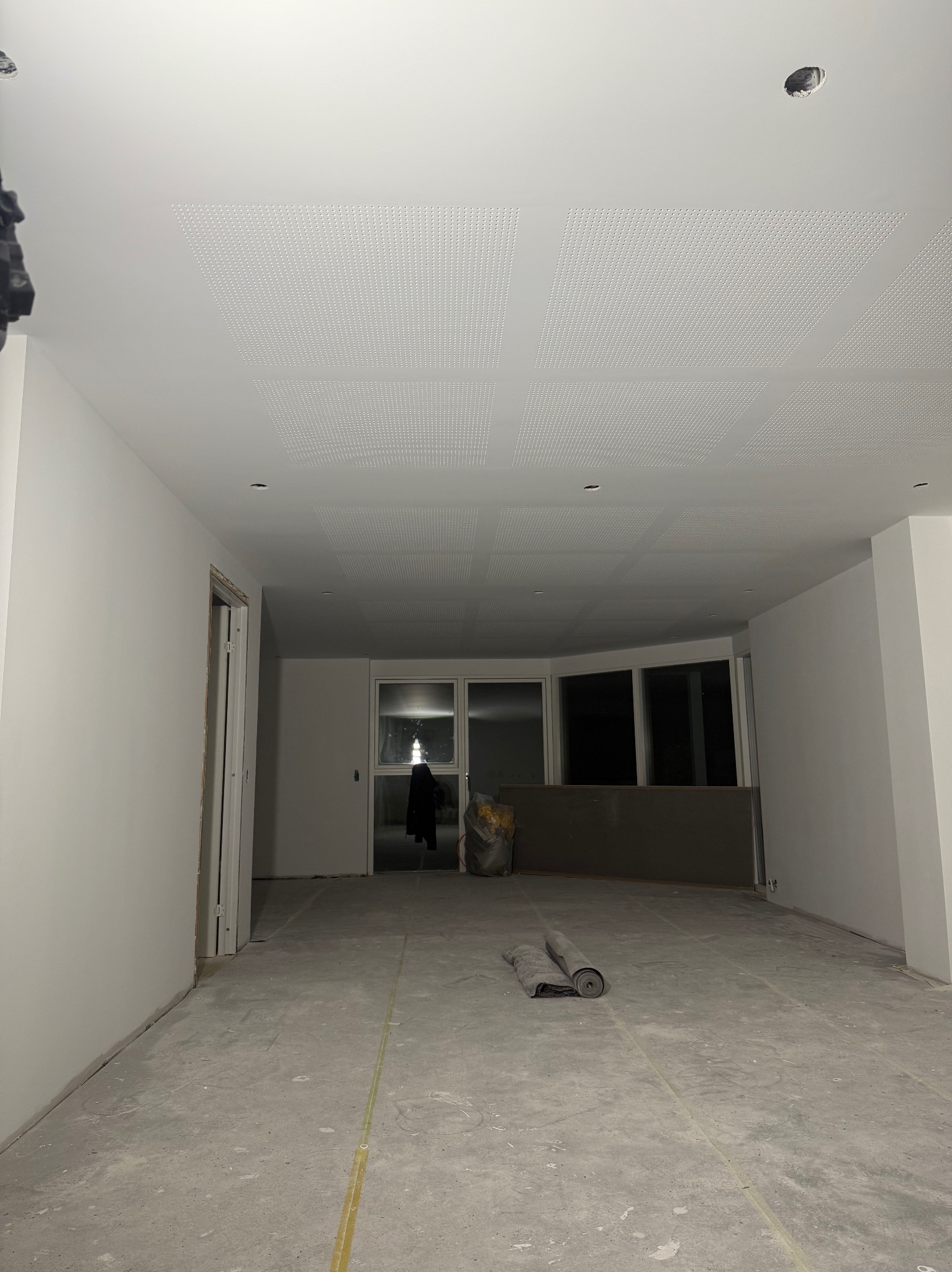 Wnętrze w trakcie prac wykończeniowych: białe ściany, sufit z perforowanymi panelami, widoczne rolki materiału na podłodze i przygotowane otwory na oświetlenie.