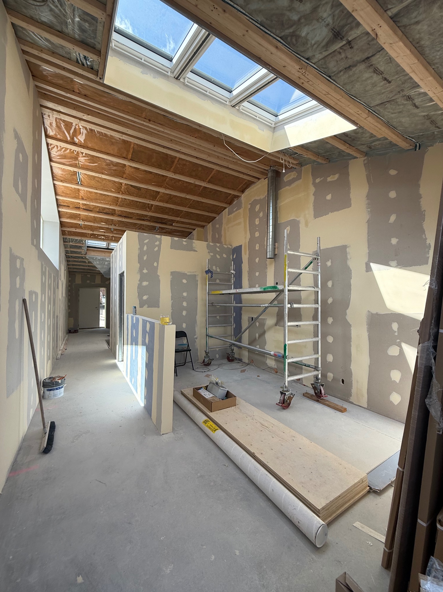 Wnętrze w trakcie prac wykończeniowych: ściany z fragmentami szpachli, rusztowanie, płyty gipsowe, okna dachowe. Widok na korytarz z otwartymi drzwiami.