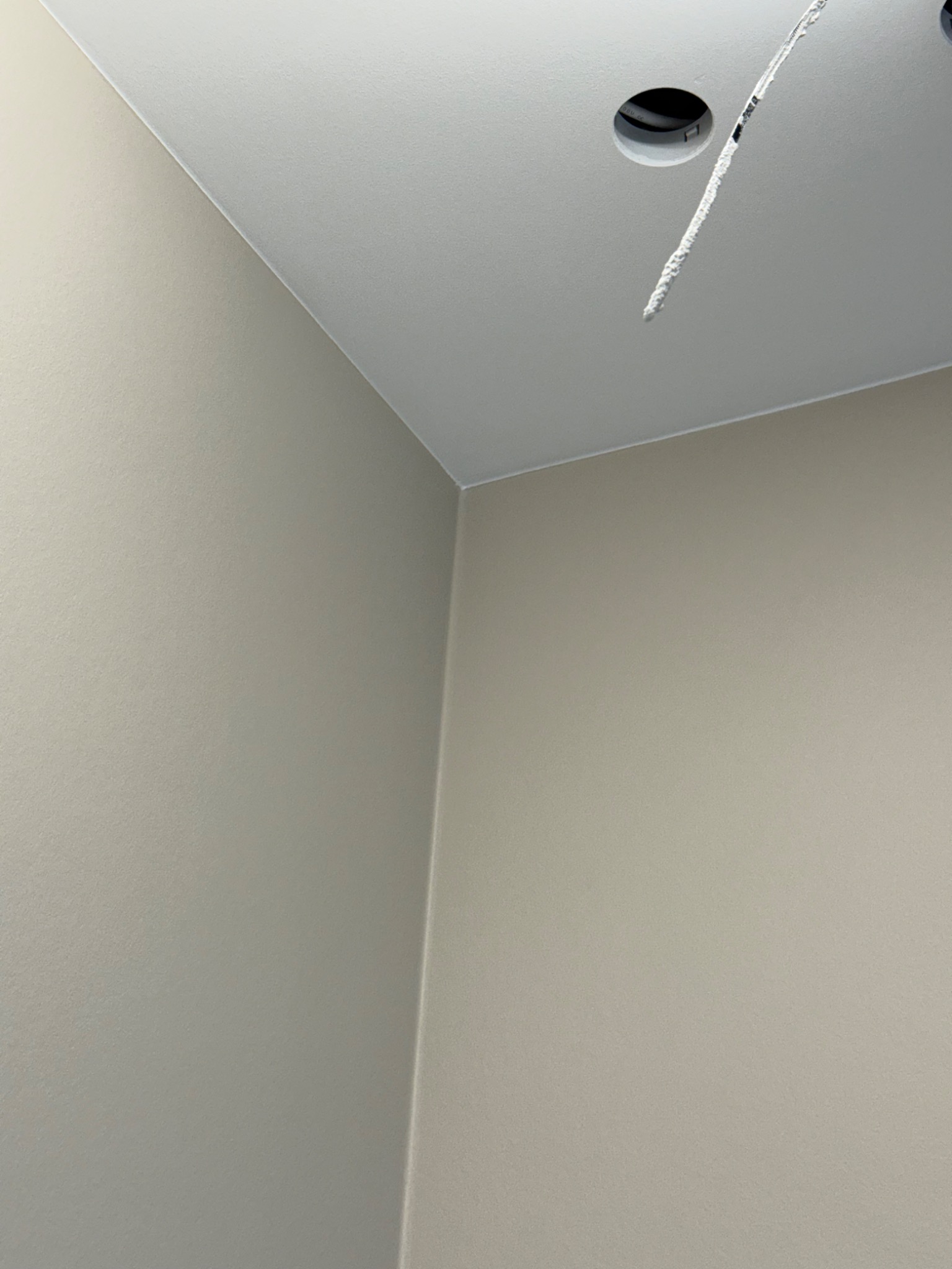 Jasnoszara, gładka ściana i sufit po malowaniu, z widocznym otworem na oświetlenie i wystającym przewodem. Minimalistyczne wykończenie wnętrza.