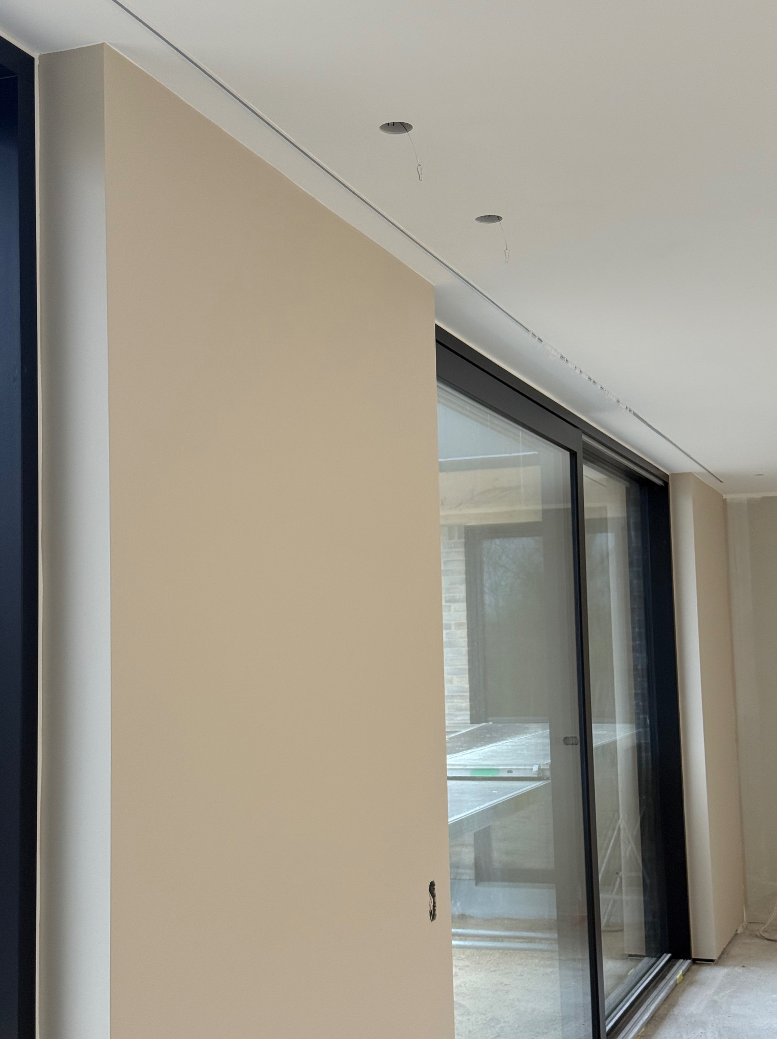 Ściana po malowaniu na beżowo, widoczne przesuwne drzwi balkonowe z czarną ramą i fragment sufitu z otworami na oświetlenie. Minimalistyczne wykończenie wnętrza.