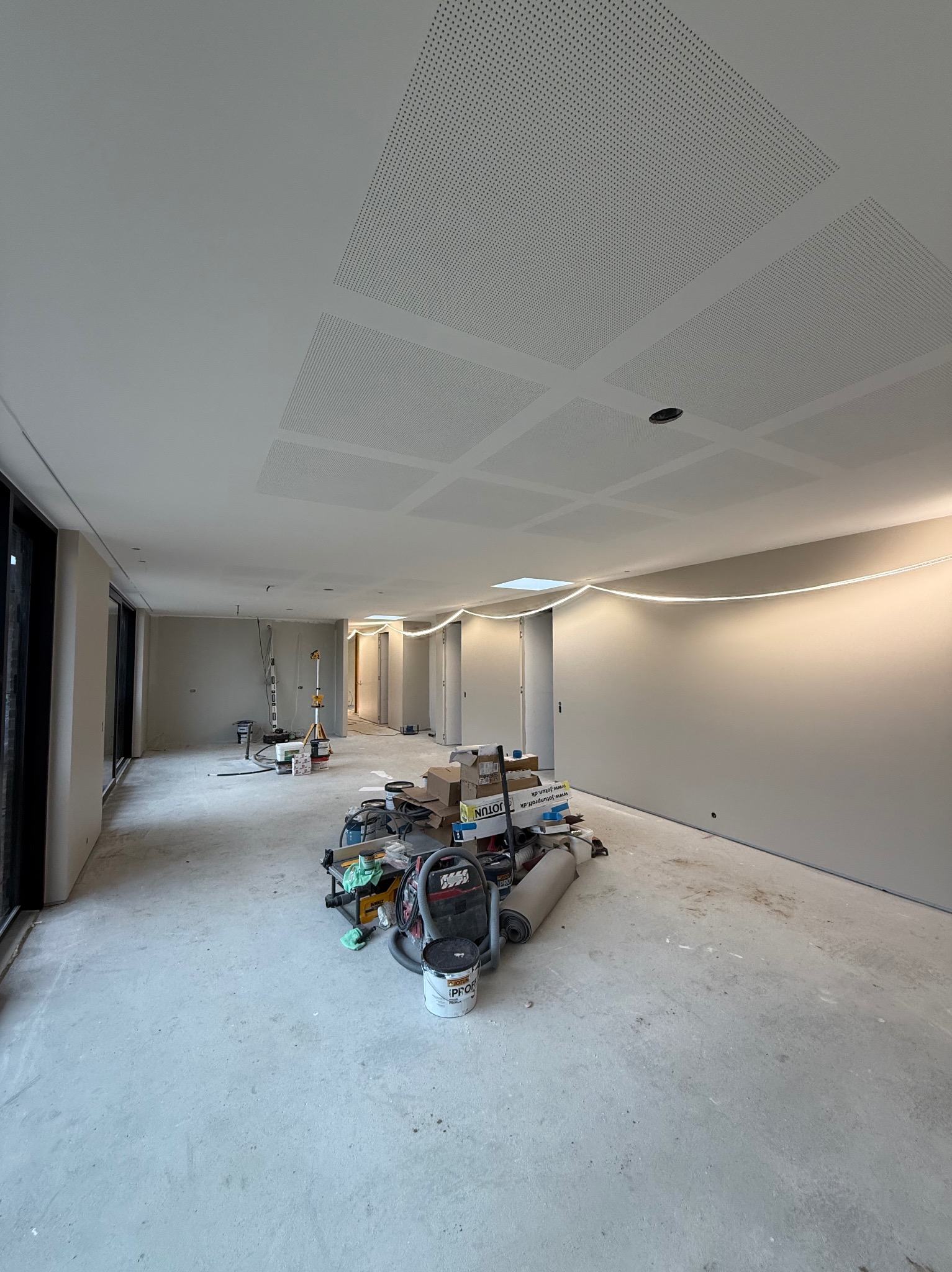 Wnętrze w trakcie prac wykończeniowych: szpachlowane ściany, sufit z perforowanymi panelami, narzędzia i materiały budowlane na podłodze. Widoczne oświetlenie LED.