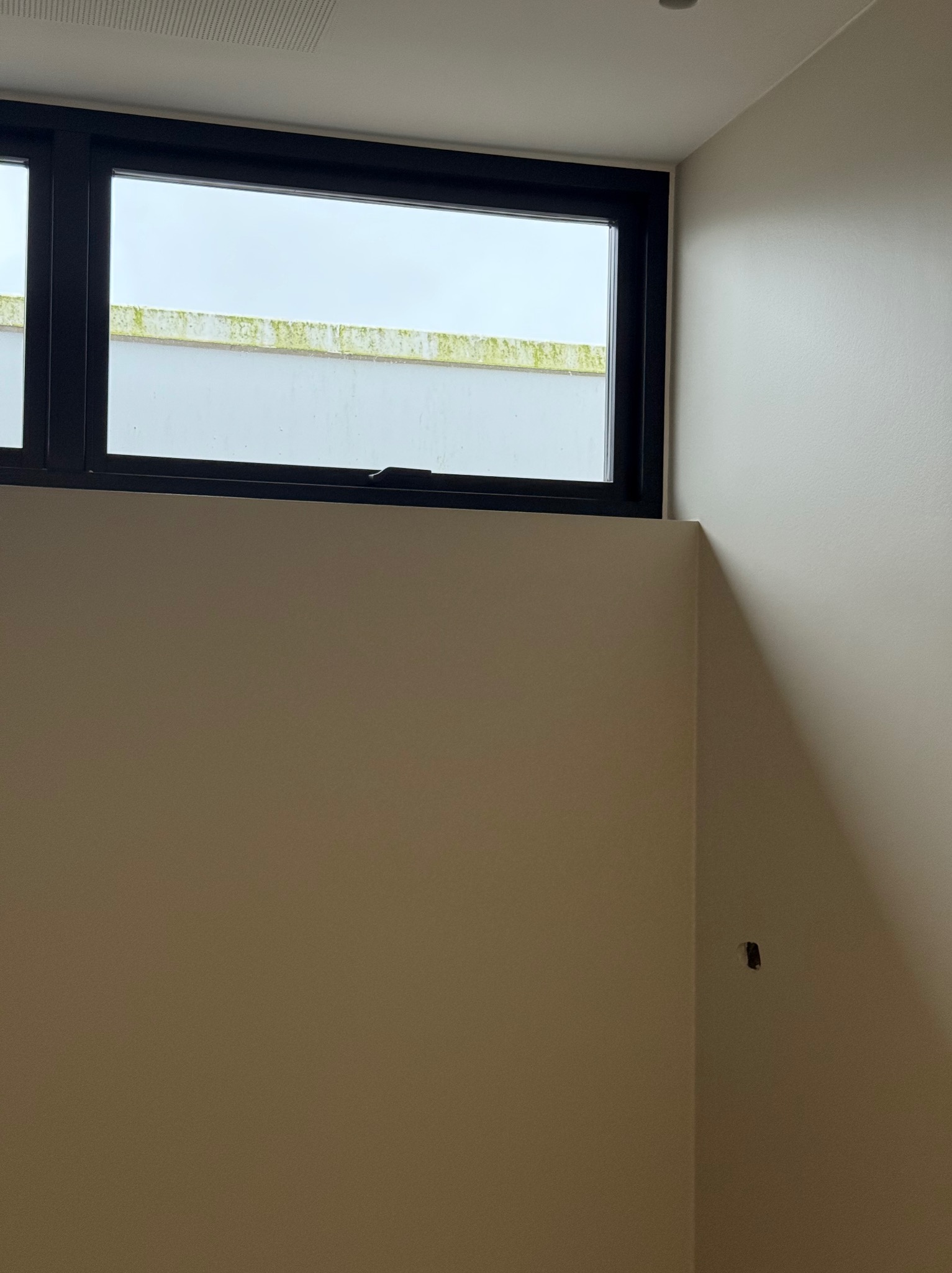 Wnętrze pokoju z oknem w czarnej ramie i gładkimi, beżowymi ścianami. Widoczny fragment ściany z otworem po gniazdku elektrycznym.