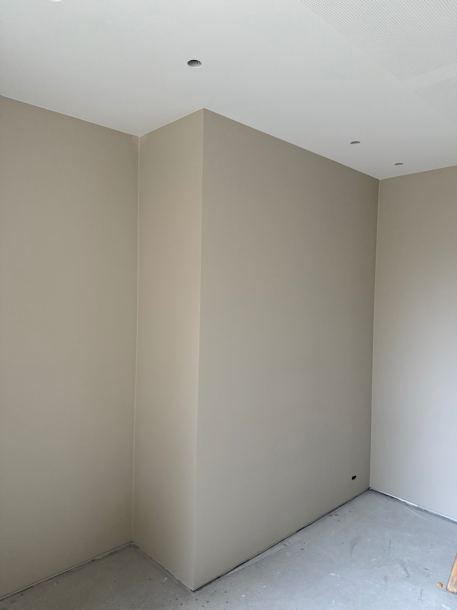 Wnętrze po malowaniu ścian na beżowo-szary odcień, widoczne gładkie powierzchnie i otwory na oświetlenie w suficie. Podłoga betonowa, sufit biały z perforacją.