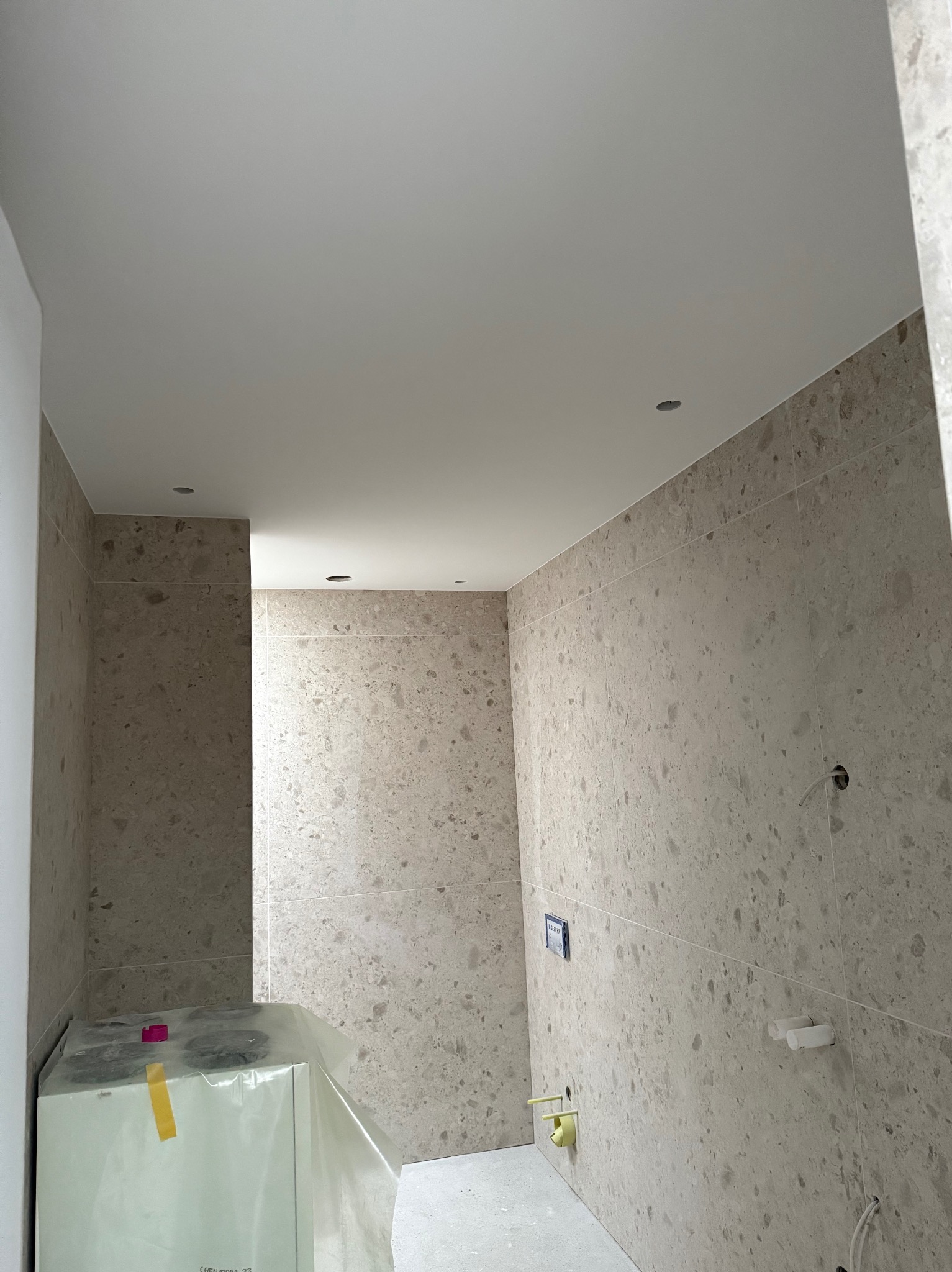 Wykończona łazienka z beżowymi płytkami imitującymi kamień, biały sufit z punktowym oświetleniem. Częściowo widoczna zabudowa stelaża WC w folii ochronnej.