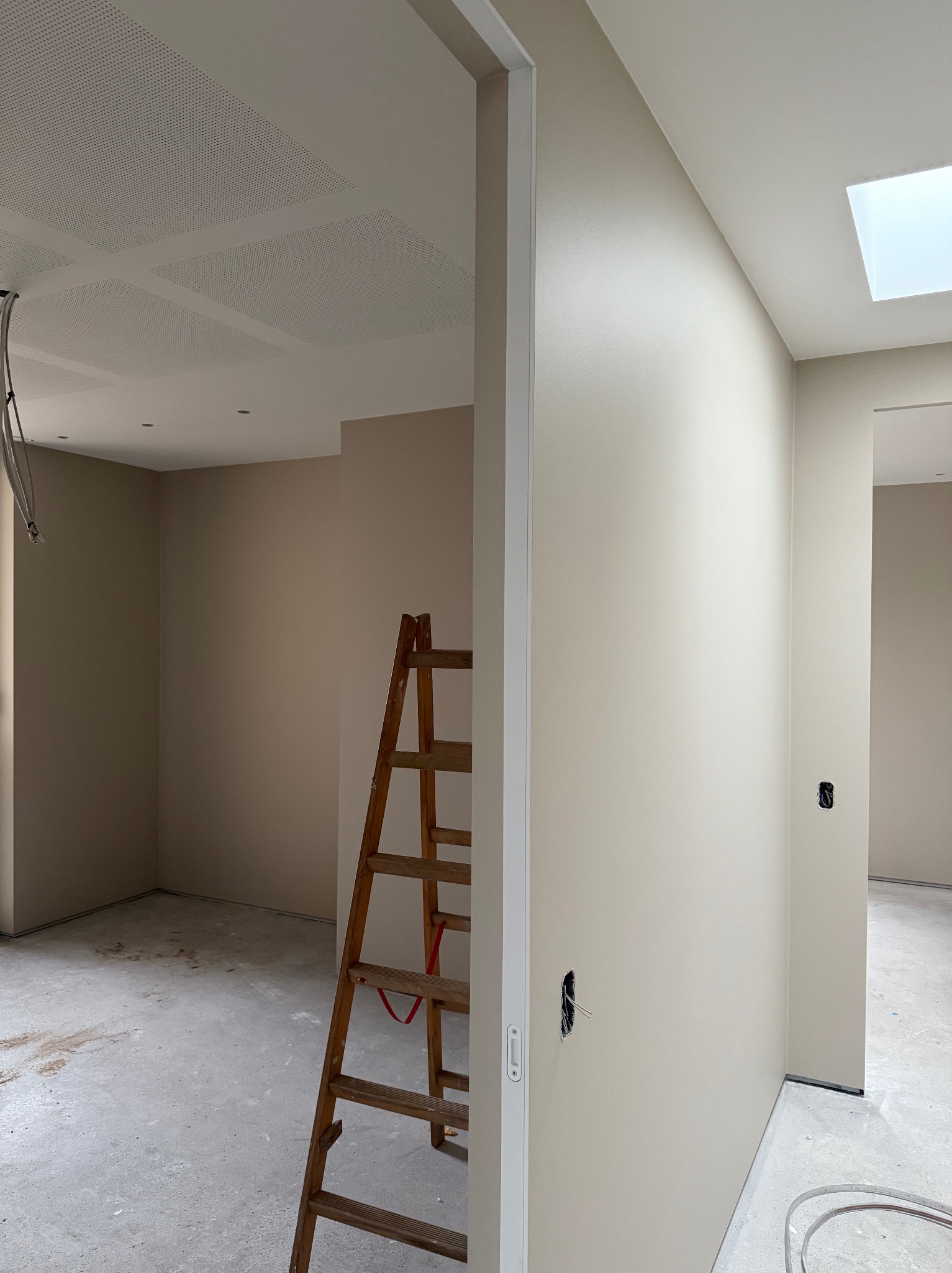 Jasne, świeżo pomalowane wnętrze z drewnianą drabiną, widoczne gniazdka elektryczne i sufit z perforowanymi panelami. W tle kolejne pomieszczenie z oknem dachowym.