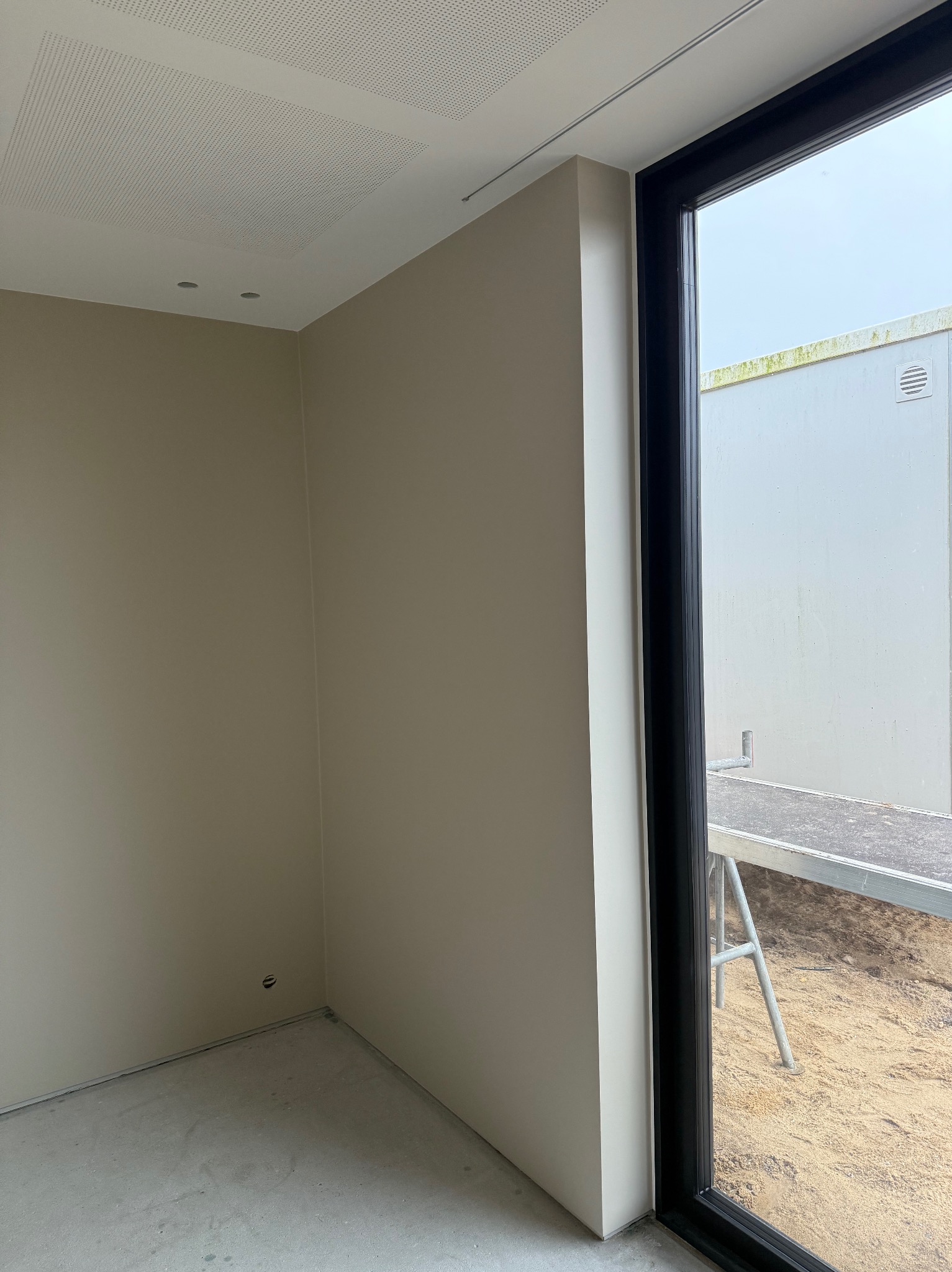 Jasne wnętrze po malowaniu ścian na beżowo, widoczny fragment sufitu z perforacją i czarna rama okienna z widokiem na zewnątrz, gdzie stoi rusztowanie.