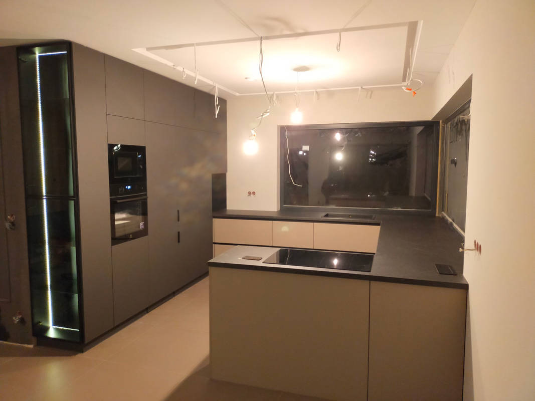 Nowoczesna kuchnia w trakcie montażu: szare szafki, wyspa z czarnym blatem, oświetlenie sufitowe w trakcie instalacji, widoczne kable i niedokończone wykończenie ścian.