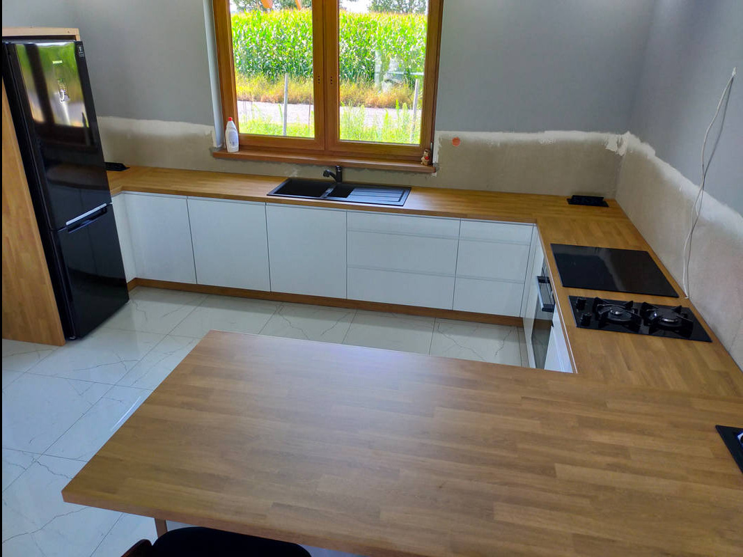 Nowoczesna kuchnia w trakcie montażu: białe szafki z drewnianym blatem, czarny zlew i płyta grzewcza, widoczne niedokończone wykończenia ścian.