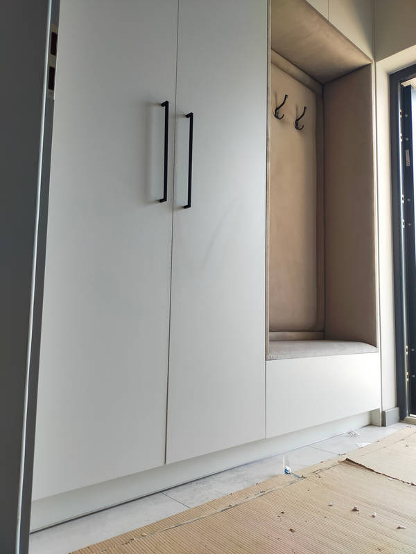 Biała szafa wnękowa z czarnymi uchwytami i wnęką z siedziskiem oraz wieszakami, widoczne zabezpieczenia podłogi.