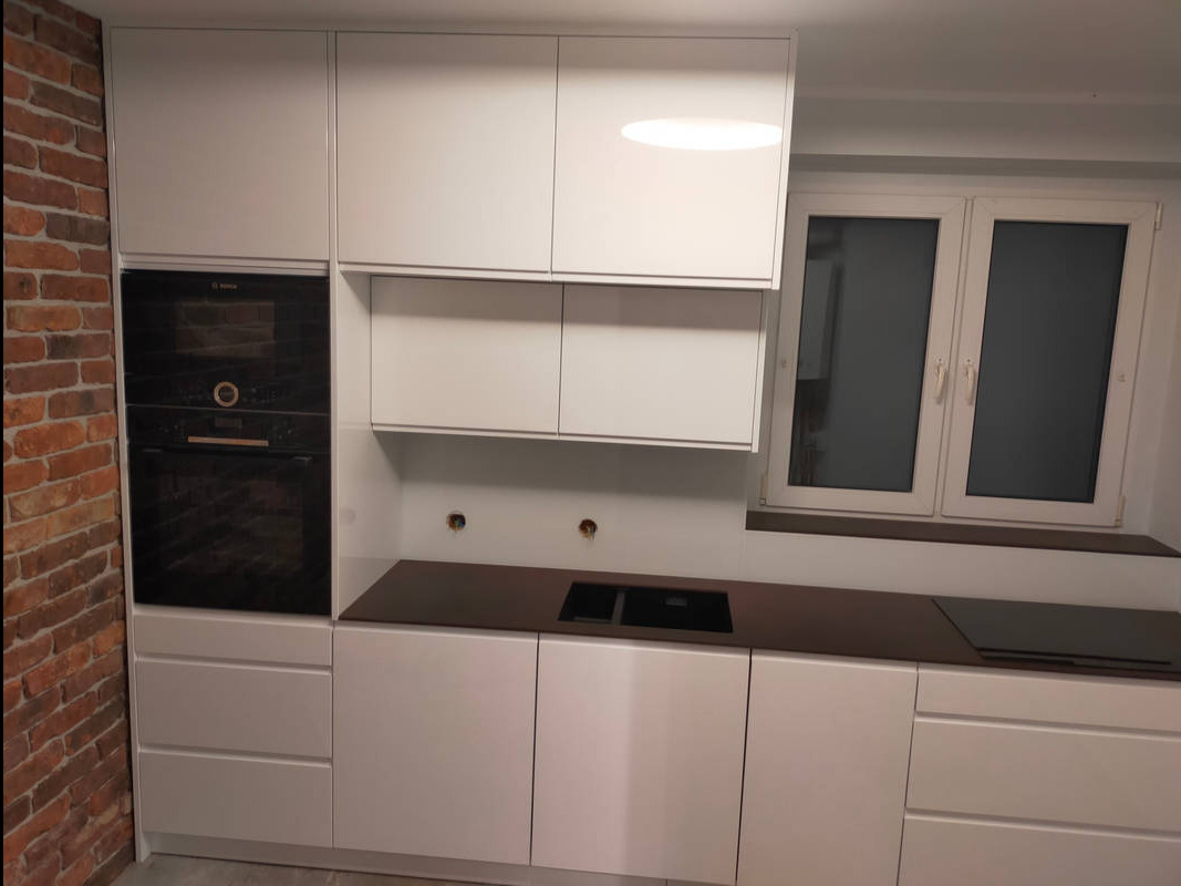 Biała, nowoczesna zabudowa kuchenna z ciemnym blatem, piekarnikiem w zabudowie i ceglaną ścianą w tle. Widoczne gniazdka elektryczne na ścianie.
