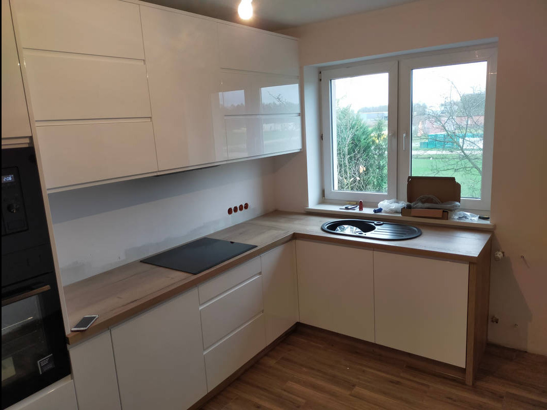 Biała kuchnia z błyszczącymi frontami, drewnianym blatem, czarną płytą grzewczą i zlewozmywakiem, widok na okno z zielenią, telefon na blacie, widoczne ślady po remoncie.