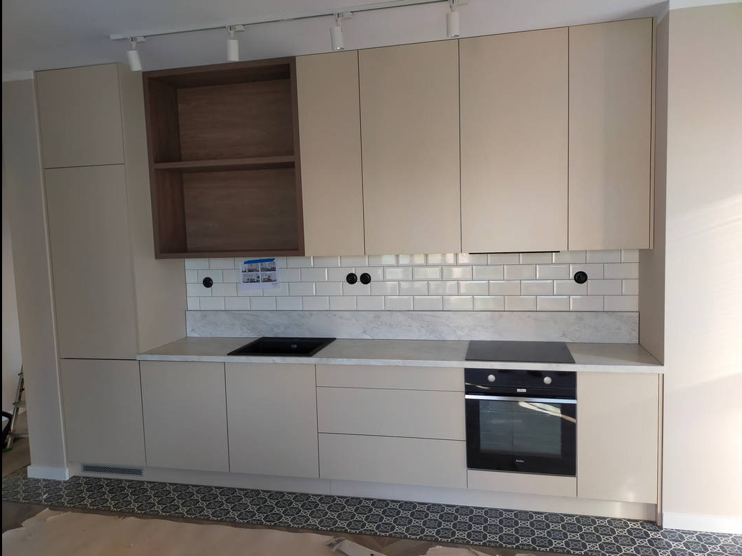 Beżowa zabudowa kuchenna z otwartą półką z ciemnego drewna, białą cegłą na ścianie i marmurowym blatem, widoczne płyty indukcyjne i piekarnik.