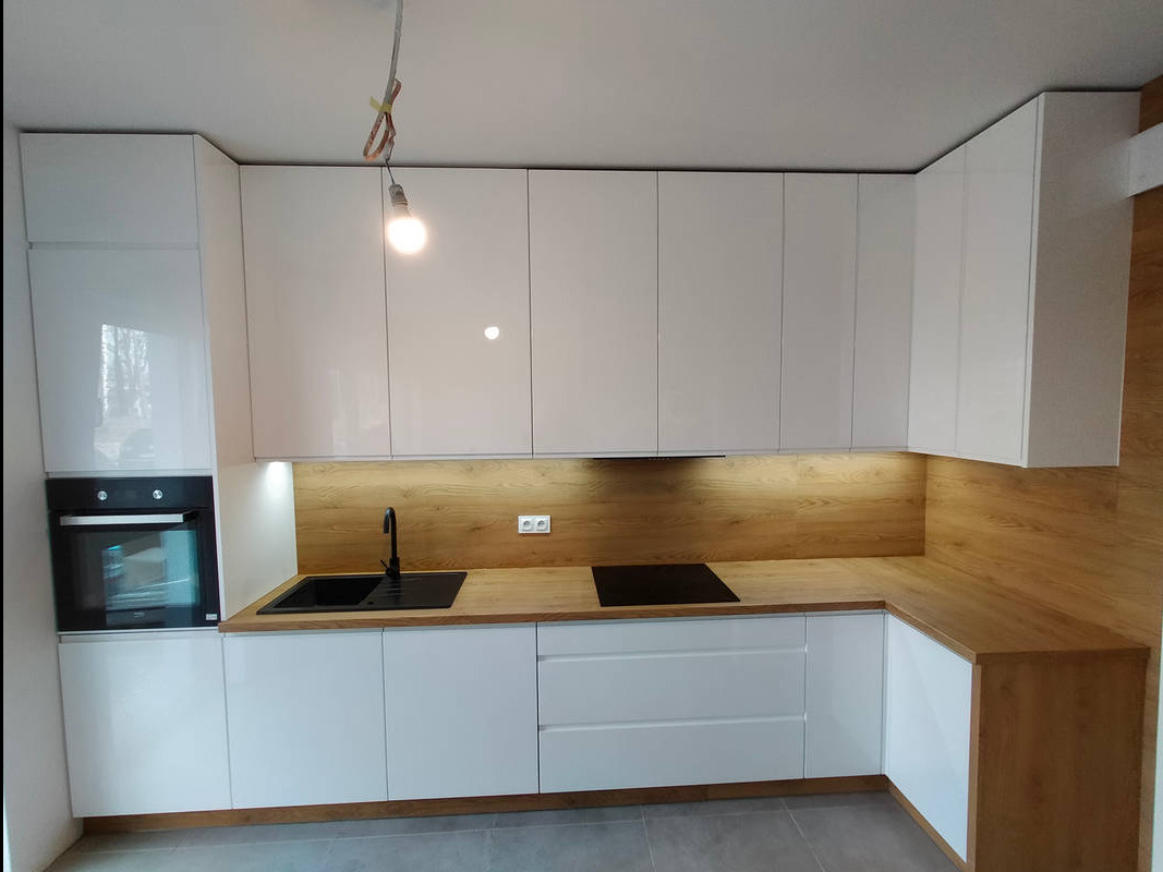 Biała, lakierowana kuchnia z drewnianym blatem i podświetleniem LED, czarny zlew i płyta indukcyjna, widoczna żarówka na kablu.