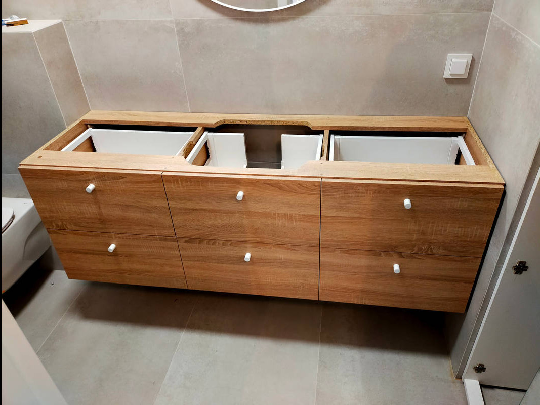 Montaż szafki łazienkowej pod umywalkę z drewnopodobnymi frontami i białymi uchwytami, widoczne białe wnętrze szuflad bez umywalki, na tle szarych płytek.