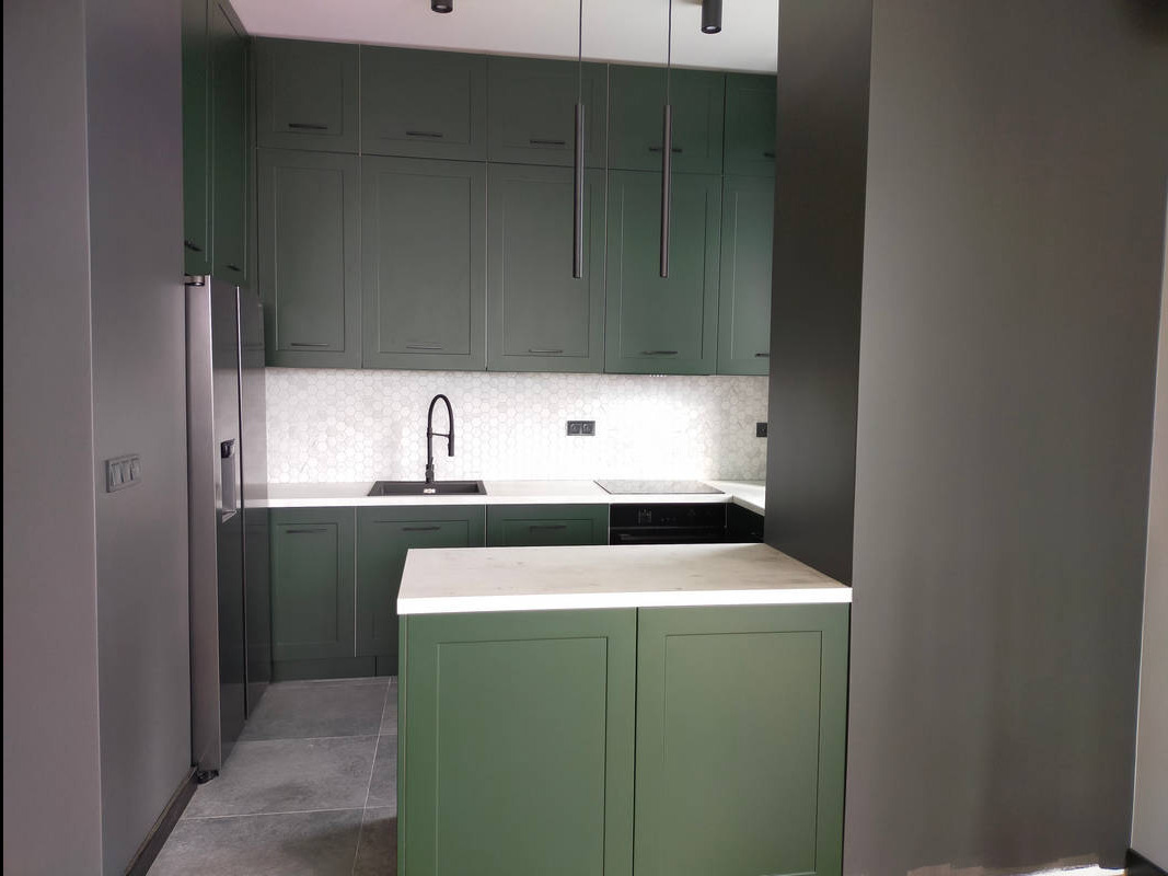 Nowoczesna kuchnia w odcieniach zieleni z wyspą, białym blatem i heksagonalnymi płytkami na ścianie.