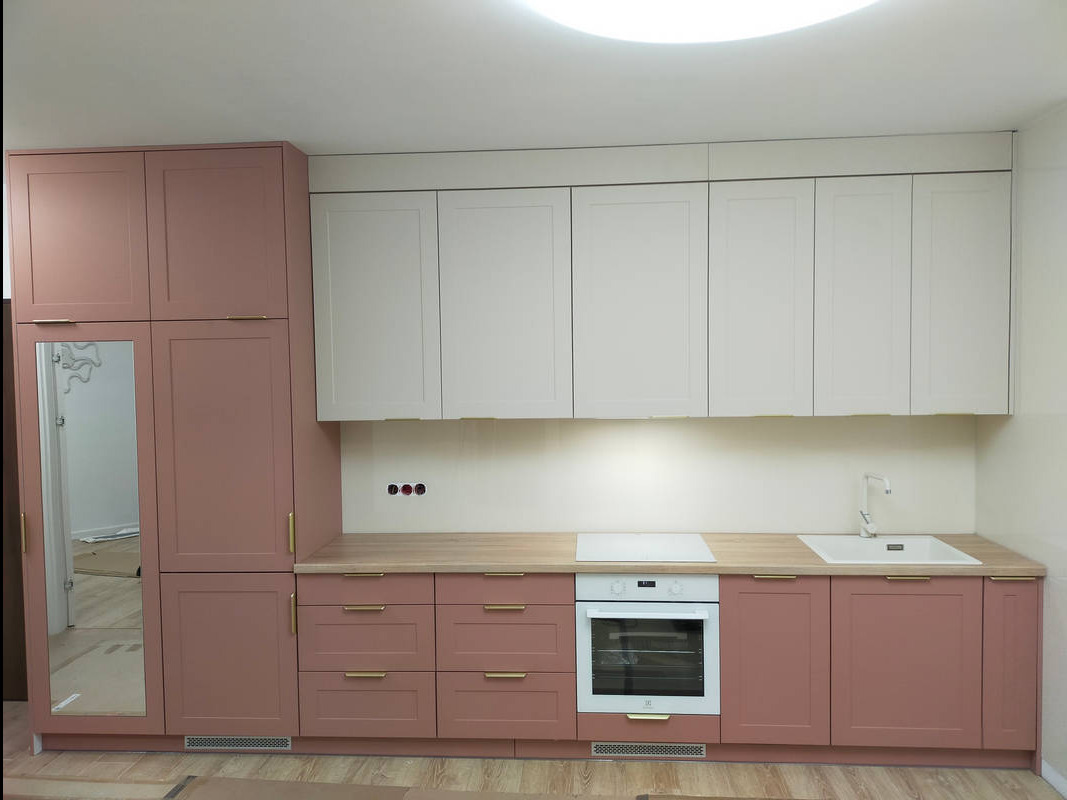 Nowoczesna kuchnia na wymiar z frontami w kolorze pudrowego różu i białymi szafkami górnymi, zintegrowany piekarnik i płyta indukcyjna, złote uchwyty, widoczny fragment podłogi w trakcie montażu.