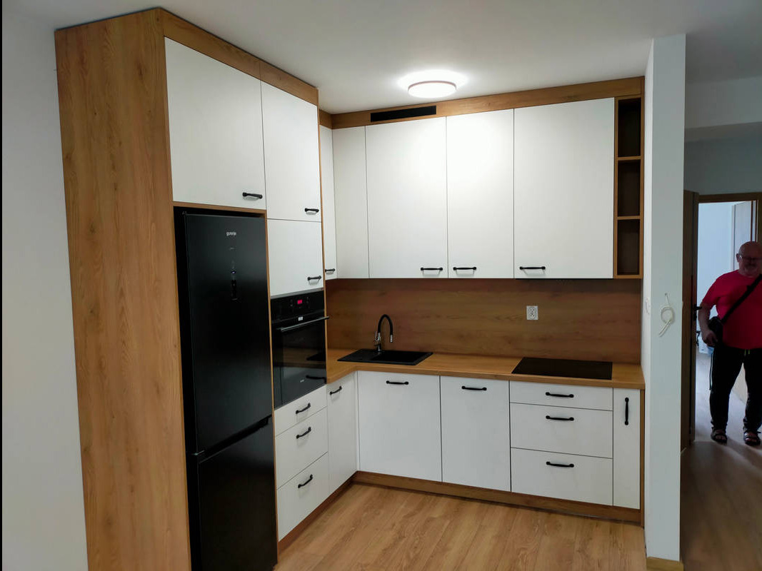 Nowoczesna kuchnia z białymi frontami i drewnianymi akcentami, wyposażona w czarny sprzęt AGD, zrealizowana w Woli Jabłońskiej. Uwagę zwraca minimalistyczny design i oświetlenie sufitowe.