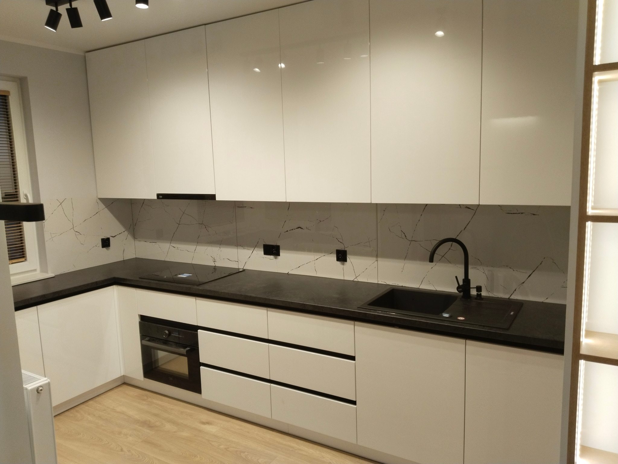 Nowoczesna kuchnia z białymi, lakierowanymi frontami szafek, czarnym blatem i czarną armaturą, wzór marmuru na ścianie, oświetlenie punktowe.