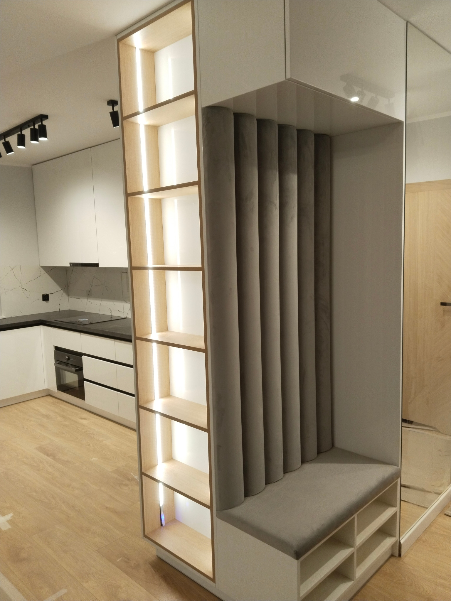 Nowoczesna kuchnia z białymi szafkami i czarnym blatem, połączona z zabudowaną szafą wnękową z półkami podświetlanymi LED i tapicerowanym siedziskiem w szarościach.