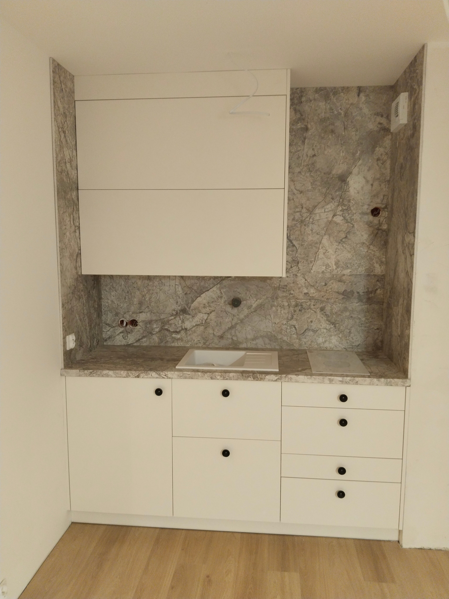 Nowoczesna, biała zabudowa kuchenna z marmurowym blatem i ścianą, widoczne gniazdka elektryczne, brak armatury przy zlewie.