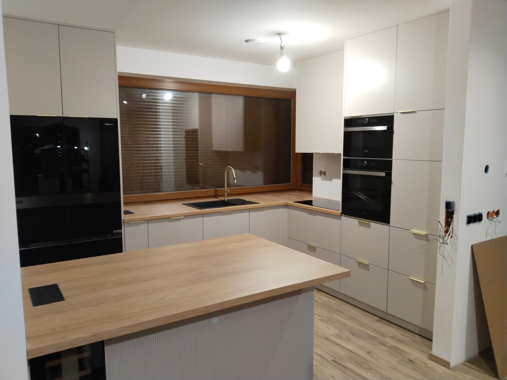 Nowoczesna kuchnia z wyspą, jasne fronty szafek, drewniany blat, czarny sprzęt AGD, okno z żaluzjami w tle, widoczne niedokończone prace elektryczne.