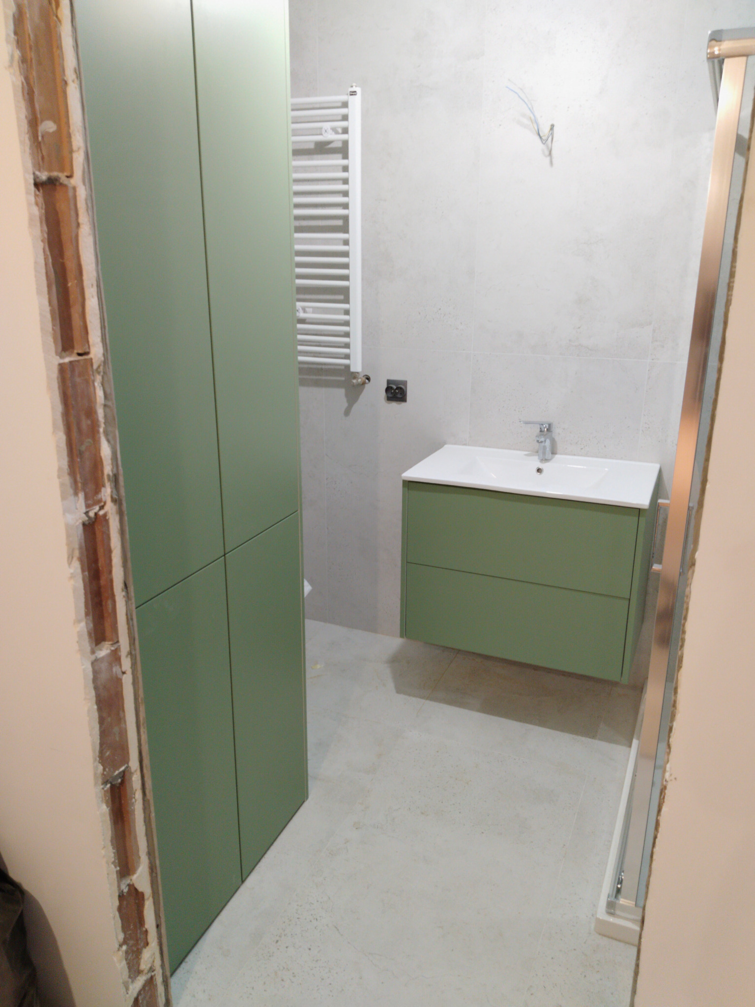 Wnętrze łazienki w trakcie remontu: widoczna zielona szafka pod umywalką, wysoka szafa w tym samym kolorze, grzejnik drabinkowy i fragment kabiny prysznicowej. Ściany w jasnych płytkach, widoczne...