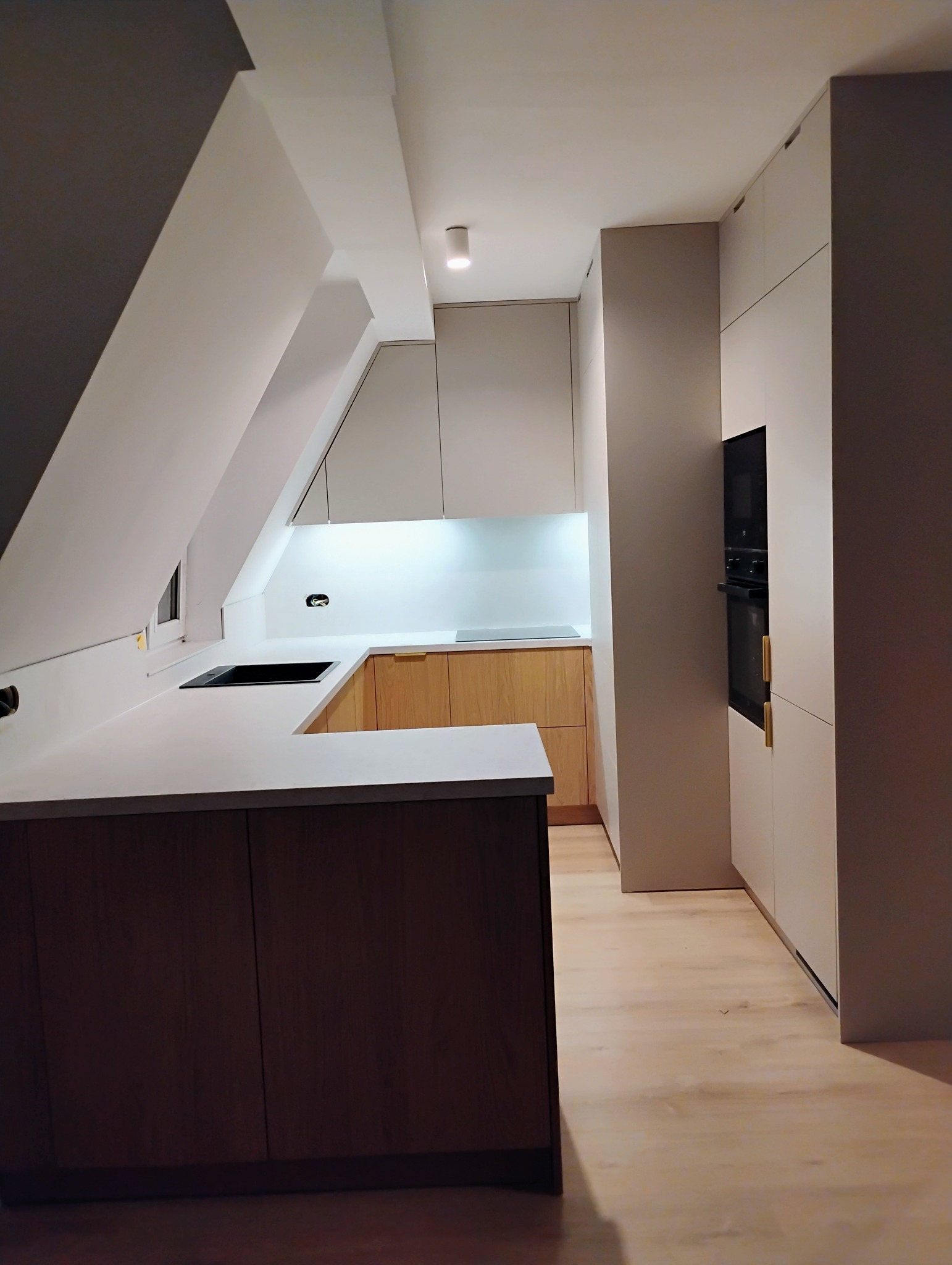 Nowoczesna kuchnia w zabudowie z jasnymi frontami, drewnianymi akcentami i białym blatem, w pomieszczeniu ze skosem.