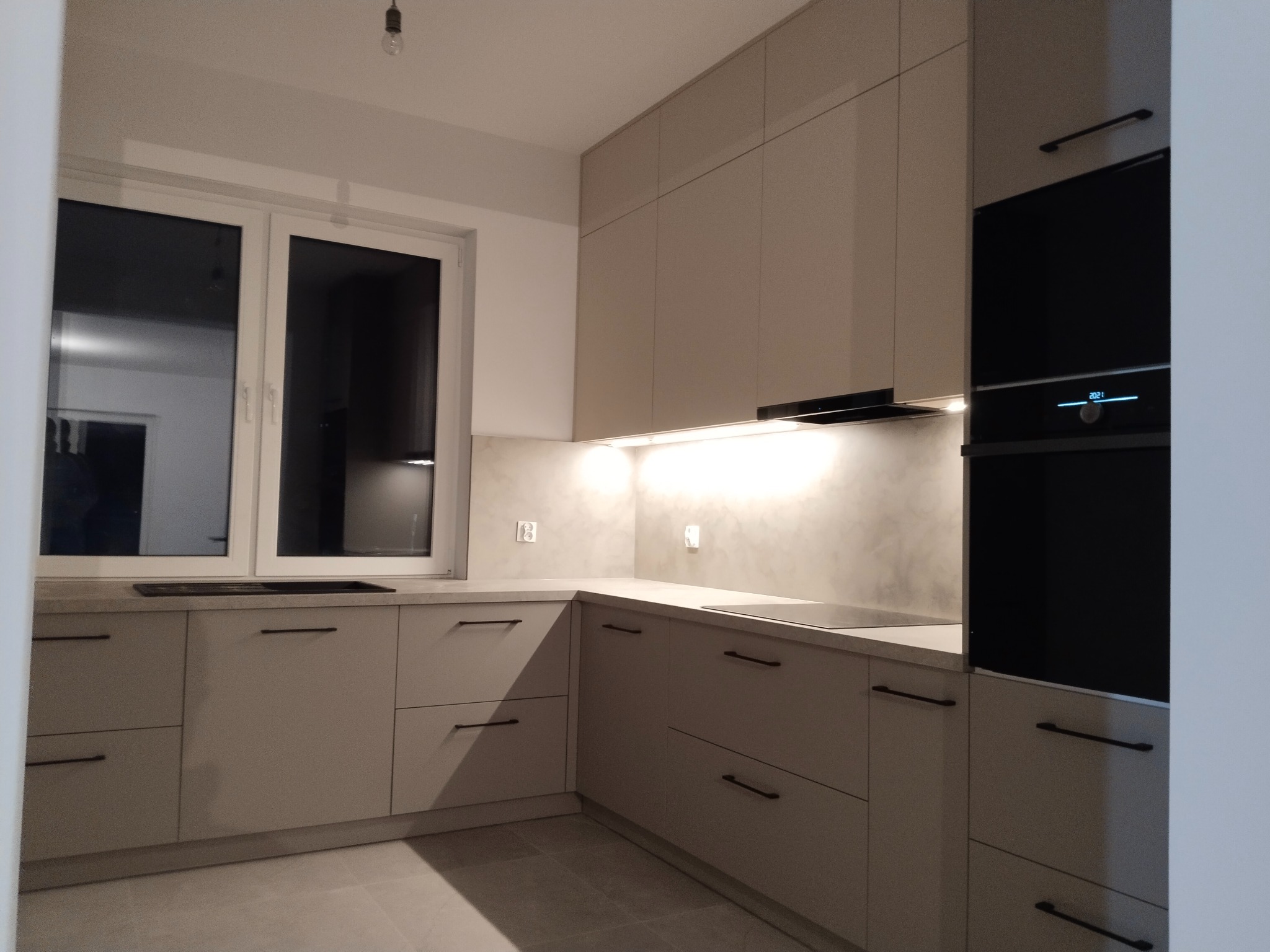 Nowoczesna, beżowa kuchnia z matowymi frontami, czarnymi uchwytami, oświetleniem LED pod szafkami wiszącymi i zabudowanym piekarnikiem.