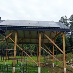Emate Łukasz Dunat - Montaż paneli na drewnianej konstrukcji z metalowym dachem, widoczne rusztowanie i przygotowany teren.