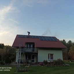 Emate Łukasz Dunat - Montaż paneli słonecznych na dachu dwukondygnacyjnego domu jednorodzinnego z drabiny, widok z dołu na tle zachmurzonego nieba i zieleni drzew.