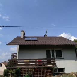 Emate Łukasz Dunat - Dom jednorodzinny z brązowym dachem, na którym zamontowane są dwa płaskie, prostokątne kolektory słoneczne. Widoczny komin, antena oraz balkon z drewnianą balustradą. Na pierwszym planie ogródek...