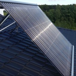 Emate Łukasz Dunat - Rząd szklanych rur kolektora słonecznego na ciemnym dachu z metalowej dachówki, wsparty na białej metalowej konstrukcji, z widocznymi czarnymi rurami izolacyjnymi, na tle zieleni drzew.