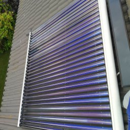 Emate Łukasz Dunat - Rząd kolektorów słonecznych z rur próżniowych, zamontowanych na szarym dachu z dachówki ceramicznej, widok z góry, z fragmentem zieleni w tle.