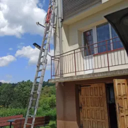 Długa aluminiowa drabina oparta o ścianę budynku z zamontowanymi roletami zewnętrznymi, widoczny fragment dachu, okno z balkonem, otwarte drewniane drzwi wejściowe oraz zieleń w tle.