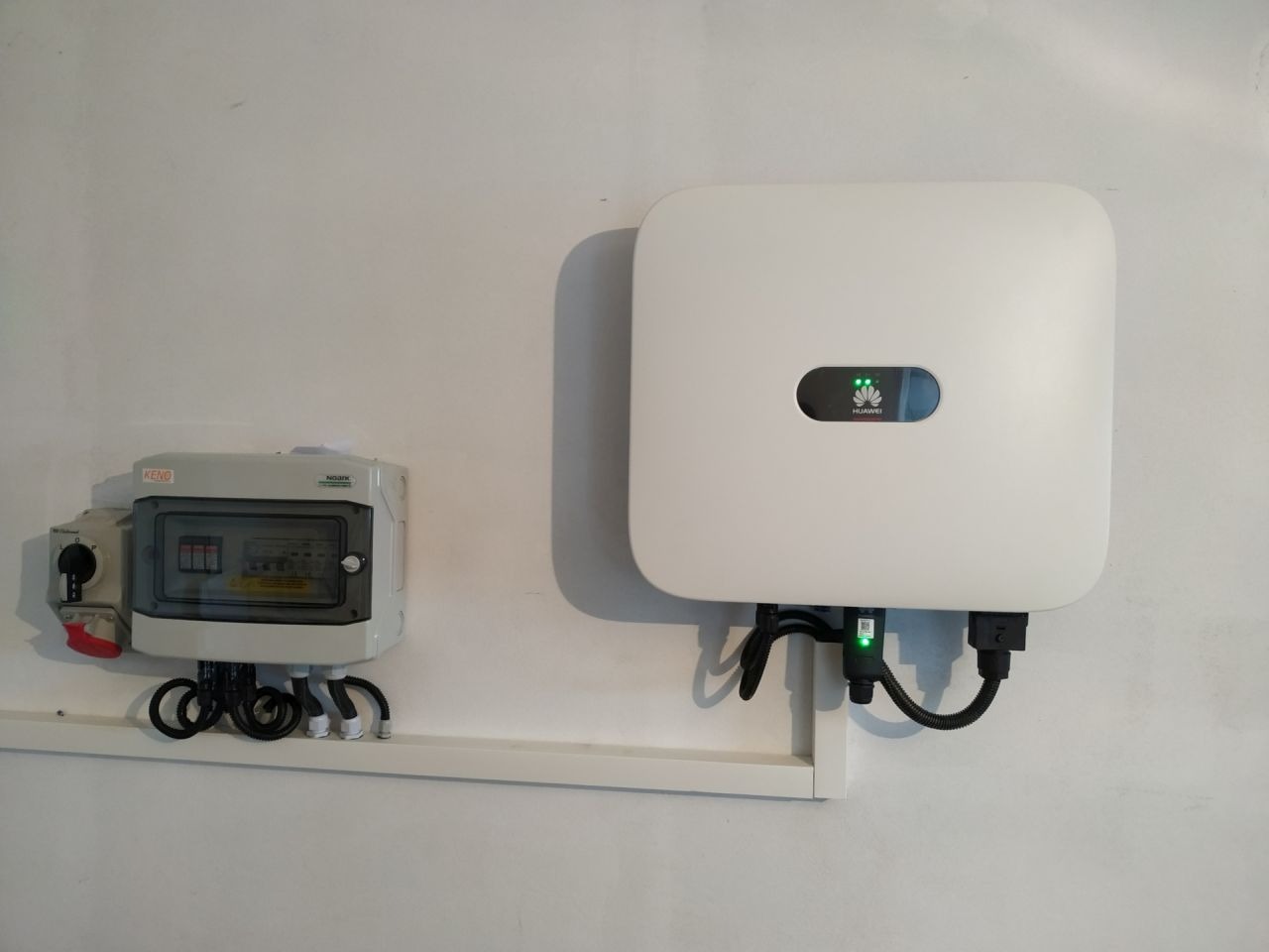 Biały inwerter Huawei z zielonymi diodami LED, zamontowany na ścianie obok szarej skrzynki bezpiecznikowej KENO z widocznymi przewodami.