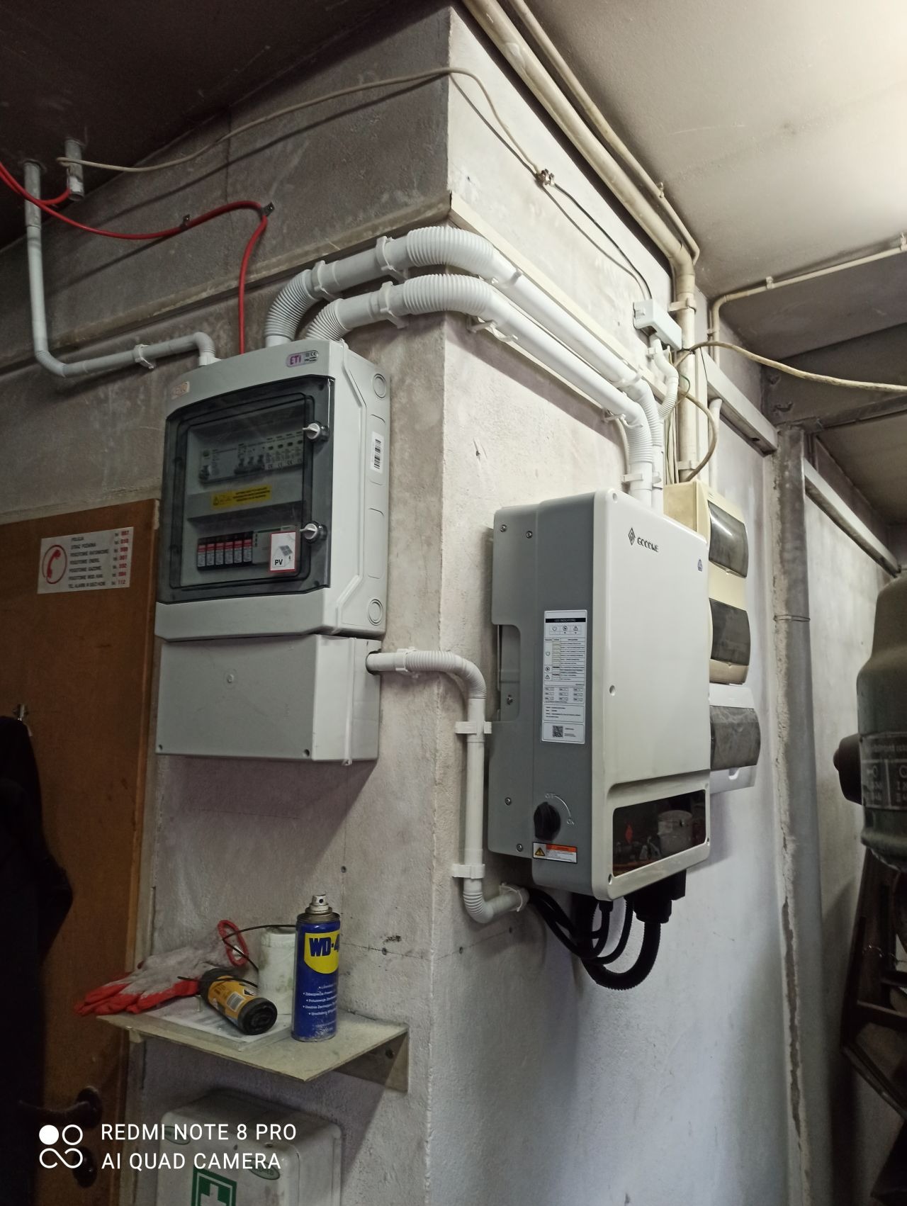 Szara skrzynka rozdzielcza fotowoltaiczna ETI i falownik GoodWe zamontowane na betonowej ścianie w piwnicy, z widocznymi przewodami w białych peszlach i narzędziami na półce poniżej.