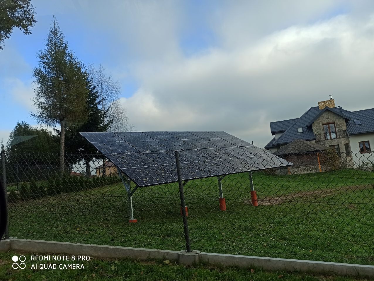 Panel słoneczny na metalowej konstrukcji z pomarańczowymi podstawami, widoczny przez siatkę ogrodzeniową na tle domu i pochmurnego nieba.