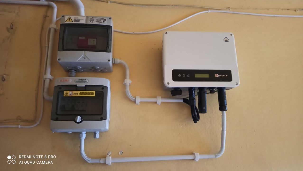 Zamontowany inwerter fotowoltaiczny GoodWe z podłączonymi przewodami i dwoma skrzynkami zabezpieczeń na żółtej ścianie.