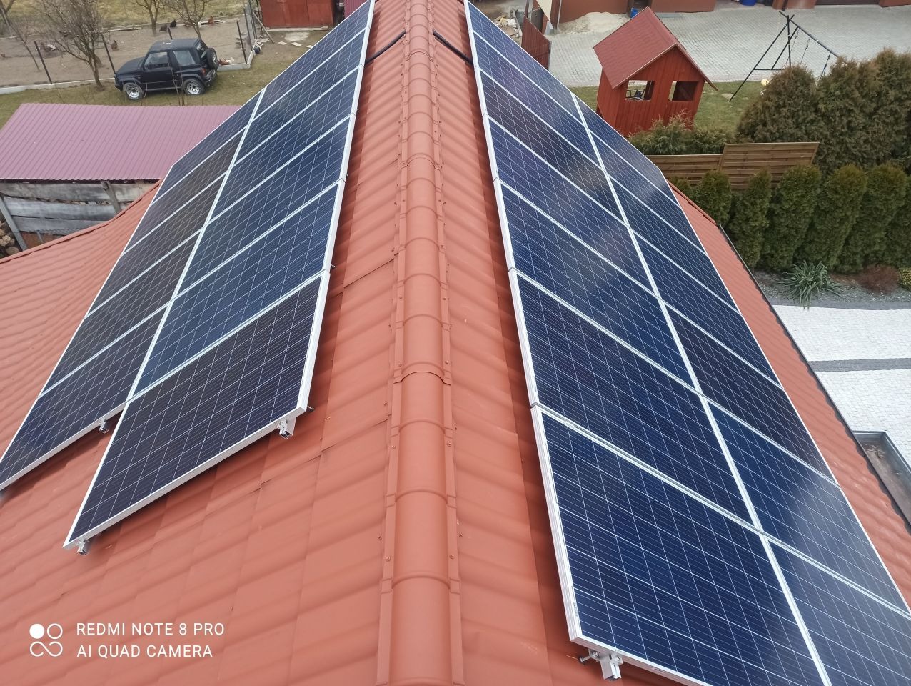 Panele słoneczne na dachu z czerwonej dachówki, widok z góry, w tle domek dla dzieci i samochód terenowy.