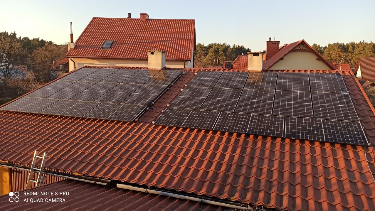 Panele słoneczne na dachu domu z czerwoną dachówką, widoczna drabina oparta o rynnę, w tle drzewa i inne budynki.