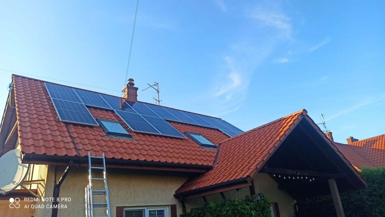 Czerwony dach z panelami słonecznymi i oknami dachowymi, widziany w słoneczny dzień. Aluminiowa drabina oparta o ścianę budynku.