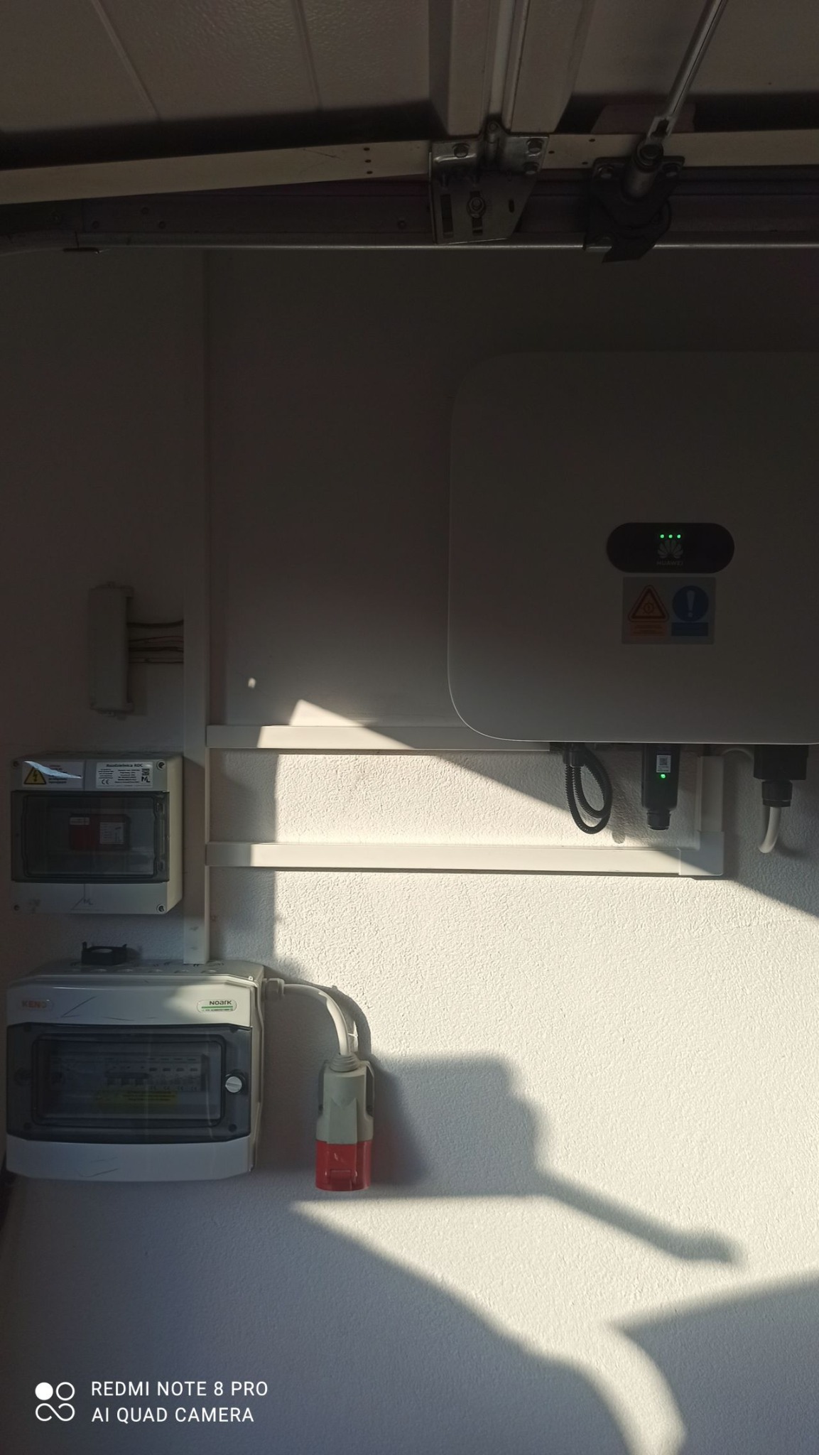 Biała ściana z zamontowaną skrzynką elektryczną, bezpiecznikami i inwerterem z diodami sygnalizacyjnymi, oświetlona popołudniowym słońcem rzucającym długi cień.